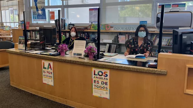 Two librarians sit behind a reference desk showcasing the LA For All campaign posters in English and Spanish