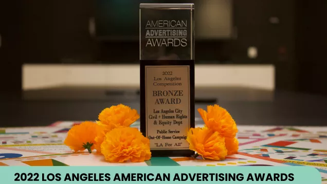 An American Advertising Award sits on top of LA for All posters beside orange flowers. Text reads "American Advertising Awards. 2022 Los Angeles Competition. Bronze Award. Los Angeles City Civil + Human Rights and Equity Depart. Public Service Out-of-home campaign. LA for All.